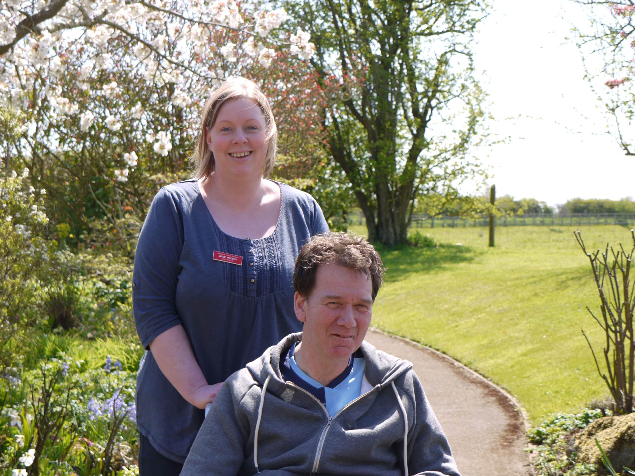 Woman pushes man in wheelchair in garden.