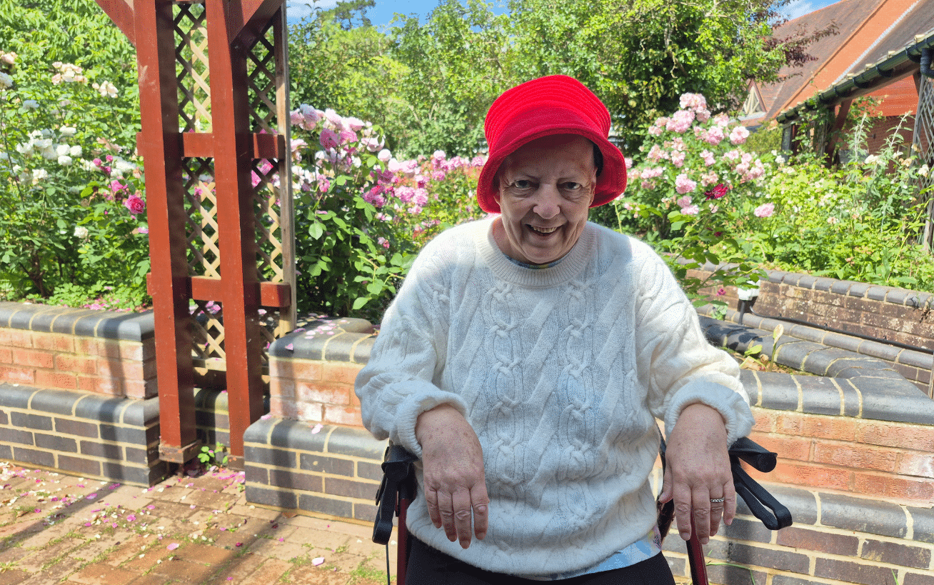 Older woman in a red hat and white sweater sits outside in a garden with flowers and greenery.