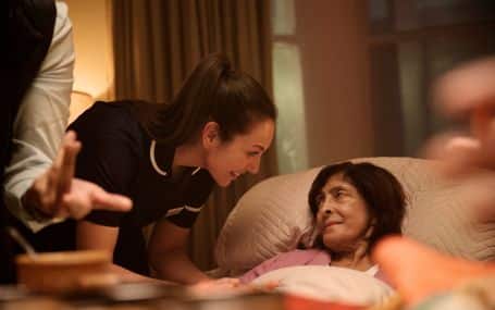 A caregiver smiles and talks to a woman resting in bed at home.