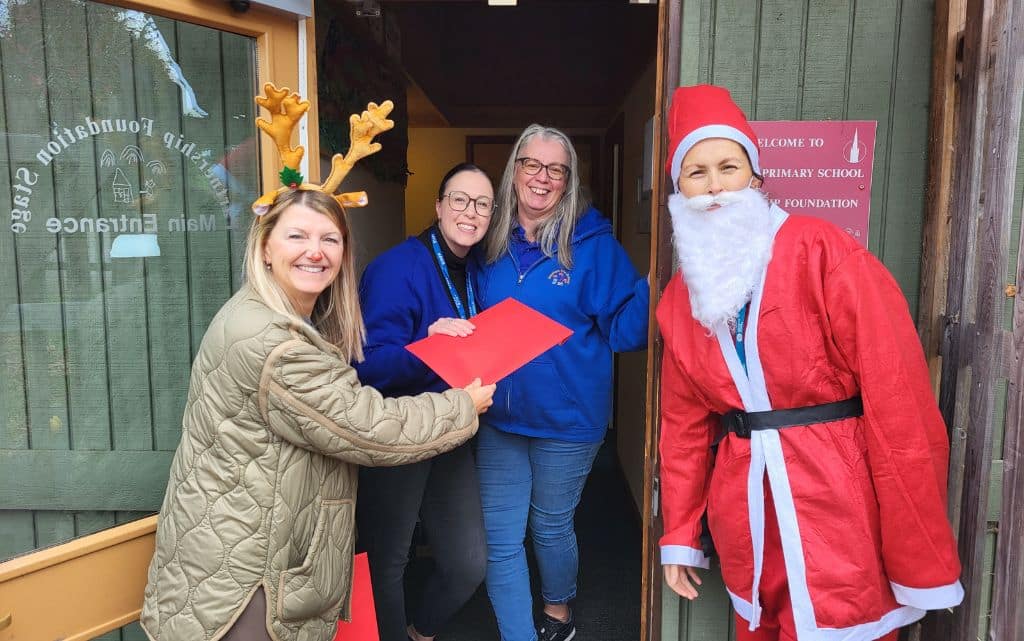 Four smiling people, one dressed as Santa, stand by a doorway holding red envelopes.